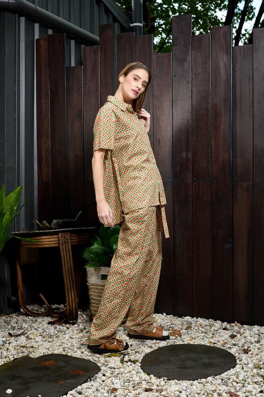 side profile of a woman wearing matching pants and shirt. The outfit is made from a soft cotton with a green and orange geo print. The pants are a straight leg relaxed fit in a high waisted tie pant, with a matching sash belt, the waistband has elastic in the back for comfort and fit and the front is in a men's suit pant style. The shirt is a short sleeve relaxed fit men's style shirt with a collar and full length hidden button placket and a straight hem that sits 3cm longer at the back.