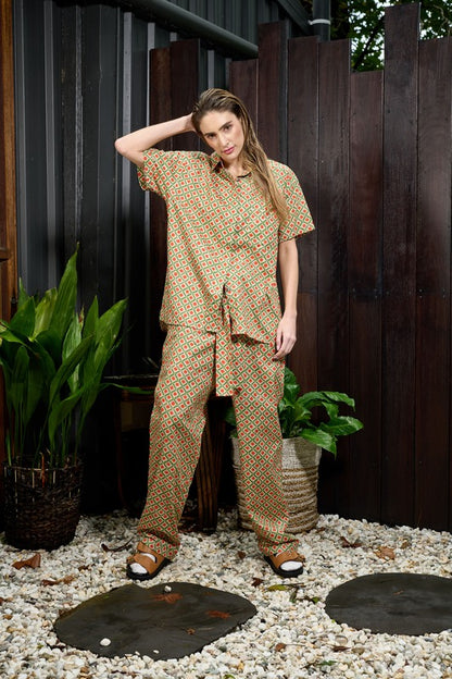 a beautiful woman wearing a chic and cool pants and shirt set, made in soft organic cotton in a green and orange geo print. The pants are soft and comfortable with elastic in the back waistband and a men's suit pant style at the front in a high waisted tie pants with a sash belt in the same fabric. The shirt is a relaxed fit short sleeve men's style with collar and full length button placket. The model is wearing chunky tan sandals to complete the fashion forward look.