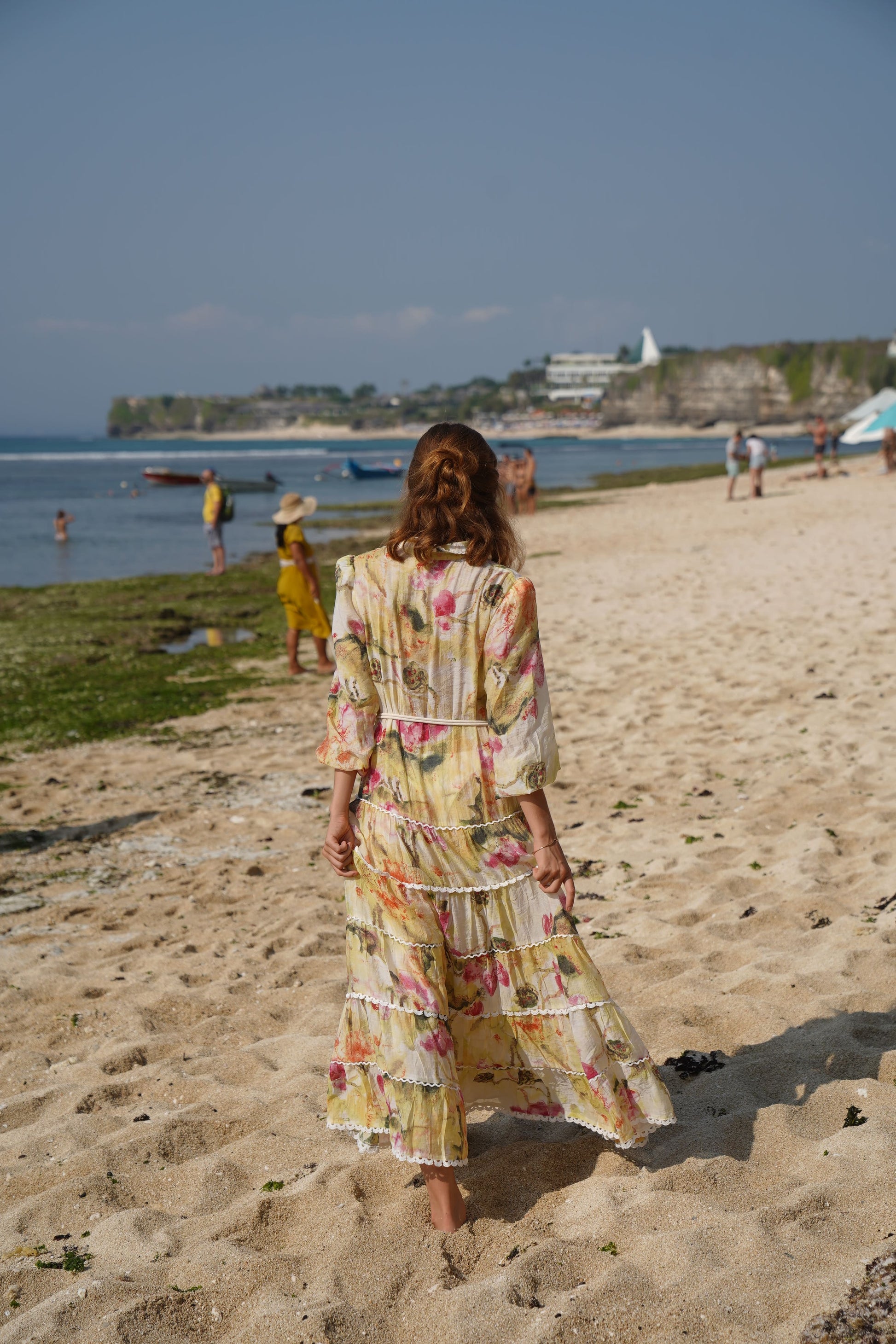 Back view of a woman on a beach wearing a relaxed fit blouson long sleeve maxi dress with a full tiered lace detailed skirt, shell button front from the drop waist, with a lace lined pea collar, on a soft lined viscose fabric with a soft white, yellow, ruby blooms print and rope belt to cinch the waist.