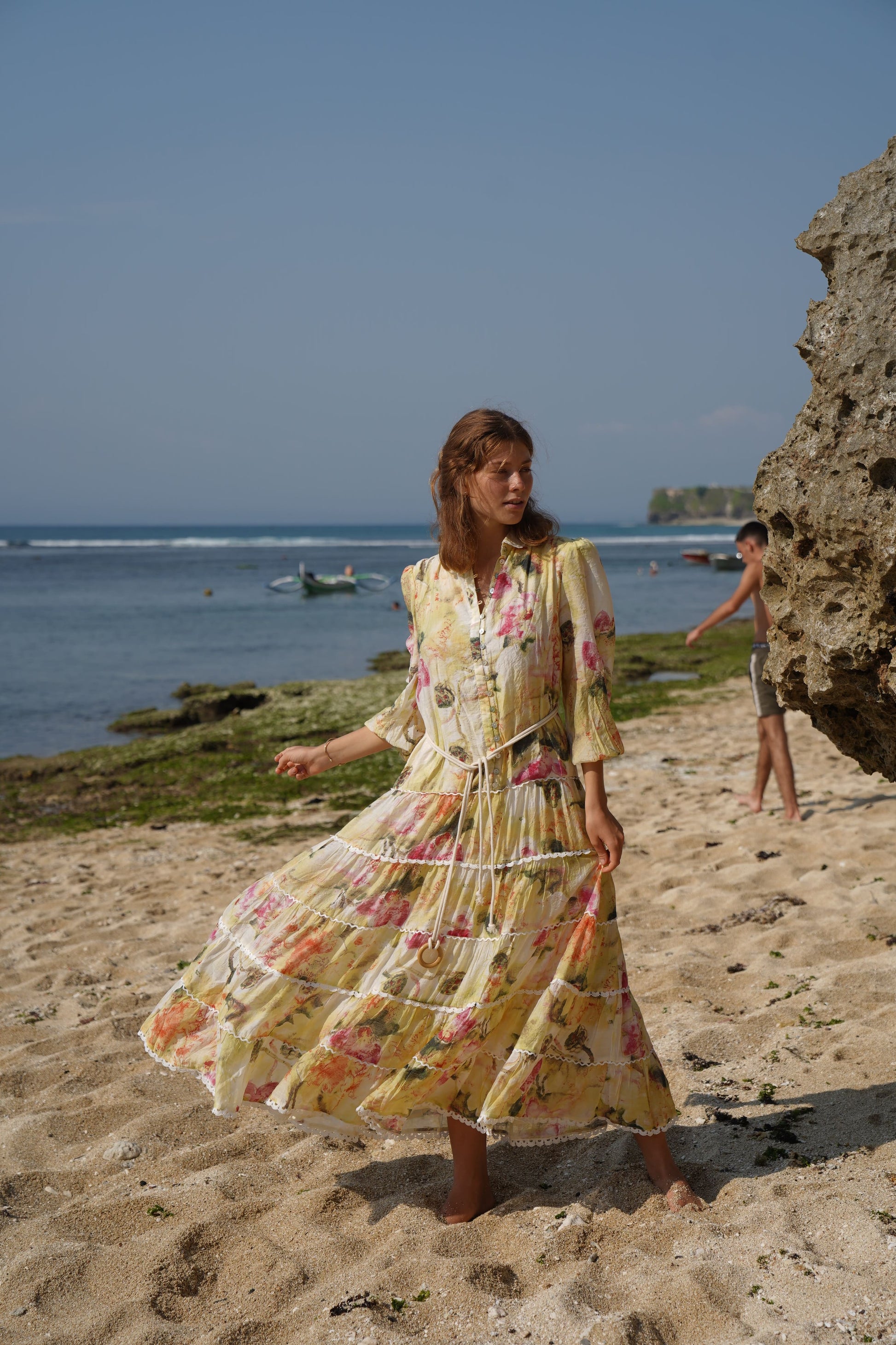 woman on a beach wearing a relaxed fit blouson long sleeve maxi dress with a full tiered lace detailed skirt, shell button front from the drop waist, with a lace lined pea collar, on a soft lined viscose fabric with a soft white, yellow, ruby blooms print and rope belt to cinch the waist.