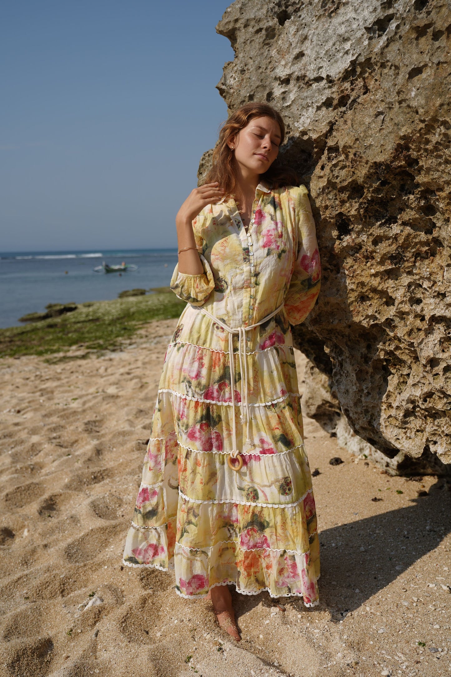 woman on a beach wearing a relaxed fit blouson long sleeve maxi dress with a full tiered lace detailed skirt, shell button front from the drop waist, with a lace lined pea collar, on a soft lined viscose fabric with a soft white, yellow, ruby blooms print and rope belt to cinch the waist.
