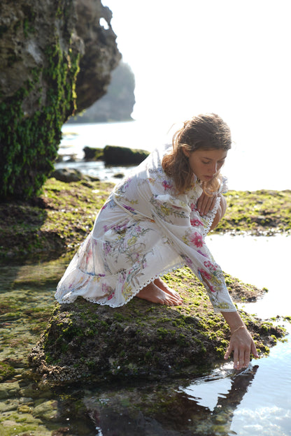 a beautiful lady crouches down on a rock with her hand stretched out to the water, she is wearing a beautiful white dress with a wildflower print in yellow, ruby, purple, pink, blue. The dress has long sheer blouson sleeves and the dress has delicate details of lace trim. The picture is romantic and whimsical.